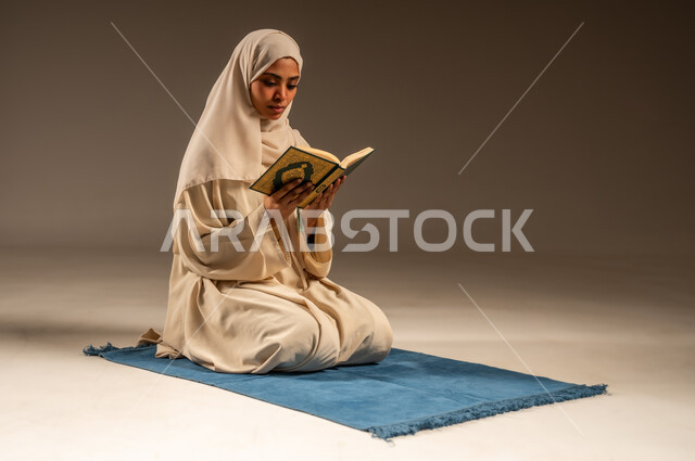 التقرب الى رب العالمين في شهر رمضان المبارك، تلاوة وقراءة كتاب الله تعالى ,الإجتهاد و الخشوع في العباده, بورتريه لامرأة عربية سعودية خليجية محجبة ترتدي العباءة تجلس على سجادة الصلاة تمسك بيدها القرآن الكريم, خلفية رمادية