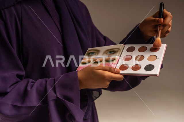 مفهوم التزين و التجمل, جودة المنتج السعودي، الاهتمام بالجمال والمظهر الخارجي, التسويق لمستحضر تجميلي نسائي, بورتريه مقرب لايدي امرأة عربية خليجية سعودية ترتدي العباءة تمسك بيدها الفرشاة وعلبة الظل, ميك اب تتوريال, خلفية رمادية