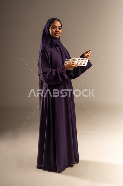 مستحضر تجميلي نسائي, ميك اب تتوريال, التسويق لمنتج سعودي, بورتريه لامرأة عربية خليجية سعودية مبتسمة محجبة ترتدي العباءة تمسك بيدها إحدى أدوات التجميل(الفرشاة) تنظر الى الكاميرا بإيماءات السرور, صورة الجسم بالطول الكامل, خلفية رمادية