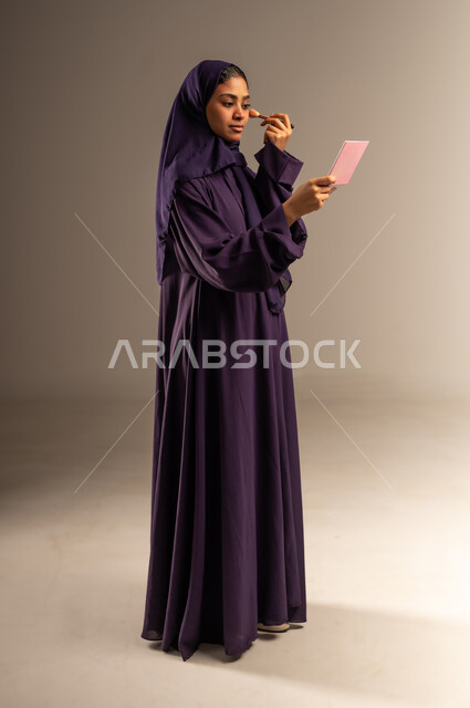 التسويق لمنتج سعودي, مستحضر تجميلي نسائي, بورتريه لامرأة عربية خليجية سعودية مبتسمة محجبة ترتدي العباءة تمسك بيدها إحدى أدوات التجميل(الفرشاة), ميك اب تتوريال, مفهوم الاهتمام بالجمال والمظهر الخارجي, صورة الجسم بالطول الكامل, خلفية رمادية