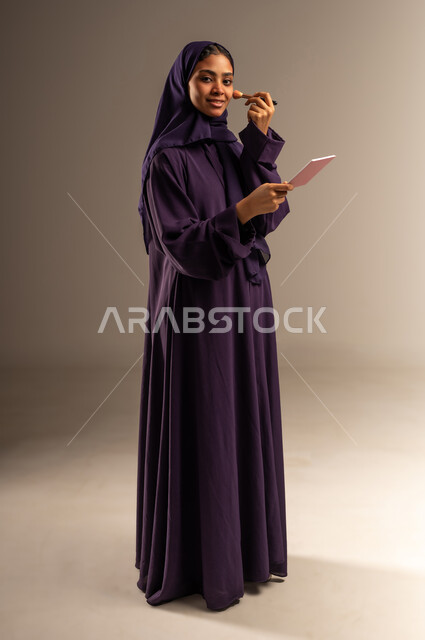 ميك اب تتوريال, التسويق لمنتج سعودي, مستحضر تجميلي نسائي, بورتريه لامرأة عربية خليجية سعودية مبتسمة محجبة ترتدي العباءة تمسك بيدها إحدى أدوات التجميل(الفرشاة) تنظر الى الكاميرا بإيماءات السرور, صورة الجسم بالطول الكامل, خلفية رمادية