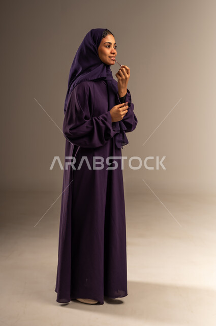جودة المنتج السعودي, مفهوم الاهتمام بالجمال والمظهر الخارجي, مستحضر تجميلي نسائي, أدوات مكياج تتوريال, بورتريه لامرأة عربية سعودية خليجية مبتسمة محجبة ترتدي العباءة تقوم بوضع أحمر الشفاه, صورة الجسم بالطول الكامل، خلفية رمادية