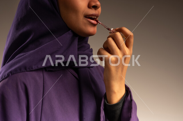 مستحضر تجميلي نسائي, جودة المنتج السعودي, مفهوم الاهتمام بالجمال والمظهر الخارجي, أدوات مكياج تتوريال, بورتريه مقرب لأيدي امرأة عربية سعودية خليجية مبتسمة محجبة تقوم بوضع أحمر الشفاه, خلفية رمادية
