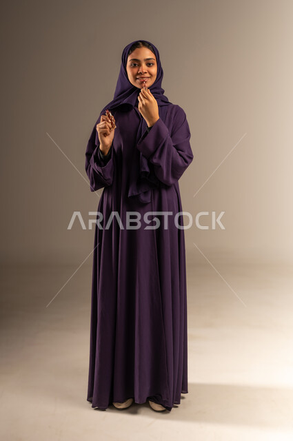 Concept of caring about beauty and appearance, women's cosmetics, Saudi product quality, makeup tutorial, portrait of a smiling veiled Saudi Arabian Gulf woman wearing an abaya applying lipstick looking at the camera with gestures of pleasure, full body image, gray background