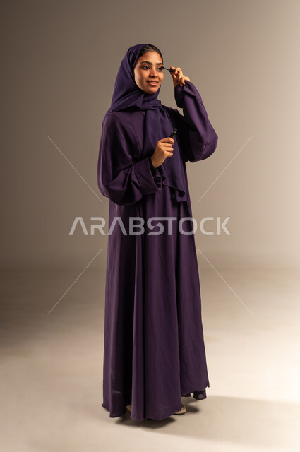 مستحضر تجميلي نسائي, جودة المنتج السعودي, أدوات مكياج تتوريال, مفهوم الاهتمام بالجمال والبشرة والمظهر الخارجي, بورتريه لامرأة عربية سعودية خليجية مبتسمة محجبة ترتدي العباءة تقوم بوضع المسكرة, صورة الجسم الطول الكامل، خلفية رمادية