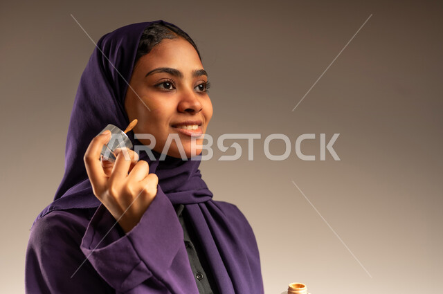 جودة المنتج السعودي, أدوات مكياج تتوريال, مفهوم الاهتمام بالجمال والبشرة والمظهر الخارجي, بورتريه مقرب لامرأة عربية سعودية خليجية مبتسمة محجبة ترتدي العباءة تمسك بيدها أدوات التجميل, مستحضر تجميلي نسائي, خلفية رمادية
