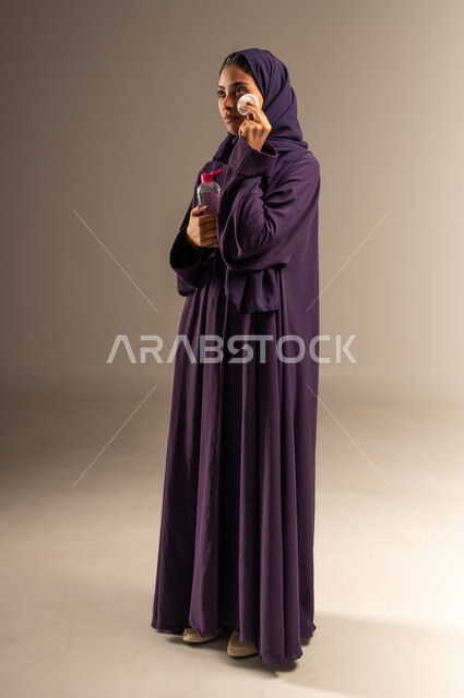 استخدام مرطب ومكون تجميلي، مفهوم العناية بنضارة وحيوية البشرة، الاهتمام بالمظهر الخارجي للنساء، بورتريه لامرأة عربية خليجية سعودية محجبة ترتدي العباءة تمسك بيدها قطنة وعبوة التونر تقوم بترطيب بشرتها، صورة الجسم بالطول الكامل، خلفية رمادية