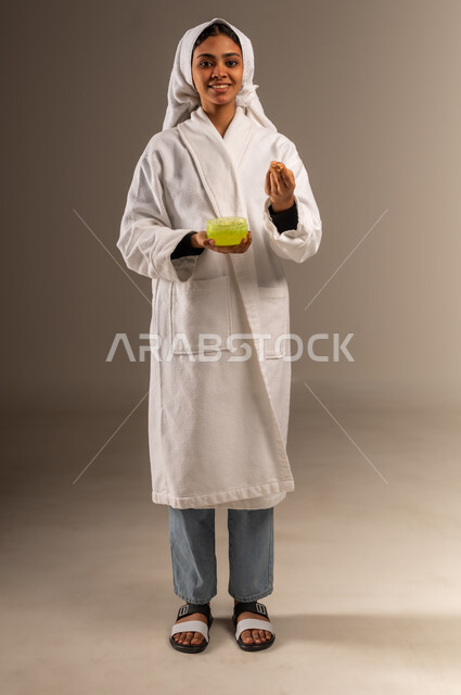 استخدام كريم ومكون تجميلي، النظر إلى الكاميرا بإيماءات السرور، مفهوم العناية بنضارة وتحسين البشرة، بورتريه لامرأة عربية خليجية سعودية مبتسمة ترتدي منشفة و روب استحمام أبيض تمسك بيدها علبة جل الترطيب، صورة الجسم بالطول الكامل، خلفية رمادية