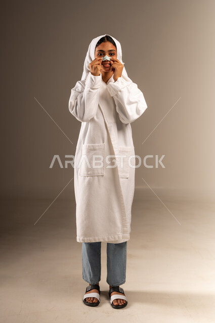 استخدام ماسك تنظيف المسام ومكون تجميلي، مفهوم العناية بنضارة وتحسين البشرة، التخلص من الرؤوس السوداء والجلد الميت، بورتريه لامرأة عربية خليجية سعودية ترتدي منشفة و روب استحمام أبيض تضع لصاقة الأنف تنظر الى الكاميرا بإيماءات السرور، صورة الجسم بالكامل، خلفية رمادية