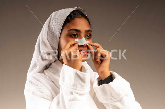 Getting rid of blackheads and dead skin, taking care of freshness and improving the skin, using a pore cleansing mask and cosmetic ingredient, close-up portrait of a Saudi Arabian Gulf woman wearing a towel and white bathrobe putting on a nose patch, the concept of caring for women's appearance, gray background