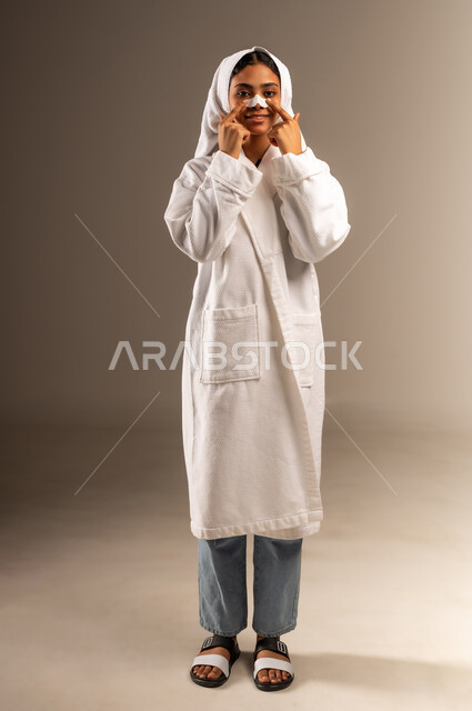 استخدام ماسك تنظيف المسام ومكون تجميلي، مفهوم العناية بنضارة وتحسين البشرة، التخلص من الرؤوس السوداء والجلد الميت، بورتريه لامرأة عربية خليجية سعودية ترتدي منشفة و روب استحمام أبيض تضع لصاقة الأنف تنظر الى الكاميرا بإيماءات السرور، صورة الجسم بالكامل، خلفية رمادية