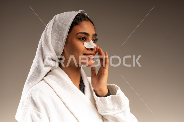 استخدام ماسك تنظيف المسام ومكون تجميلي، التخلص من الرؤوس السوداء والجلد الميت، العناية بنضارة وتحسين البشرة، بورتريه مقرب لامرأة عربية خليجية سعودية ترتدي منشفة و روب استحمام أبيض تضع لصاقة الأنف، مفهوم الاهتمام بالمظهر الخارجي للنساء، خلفية رمادية