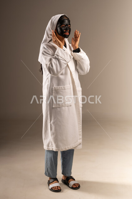 استخدام مكون  ومستحضر تجميلي، خطوات تنظيف وتقشير البشرة، مفهوم العناية والاهتمام بالمظهر الخارجي عند النساء، بورتريه لامرأة عربية خليجية سعودية ترتدي منشفة وروب الاستحمام الابيض تقوم بوضع قناع الطين على وجهها، صورة الجسم بالكامل، خلفية رمادية