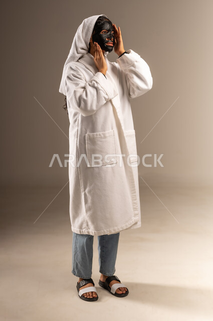 خطوات تنظيف وتقشير البشرة، مفهوم العناية والاهتمام بالمظهر الخارجي عند النساء، استخدام مكون  ومستحضر تجميلي، بورتريه لامرأة عربية خليجية سعودية ترتدي منشفة وروب الاستحمام الابيض تقوم بوضع قناع الطين على وجهها، صورة الجسم بالكامل، خلفية رمادية