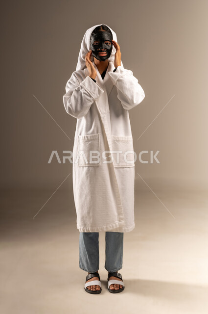 خطوات تنظيف وتقشير البشرة، النظر الى الكاميرا بإيماءات السرور، استخدام مكون ومستحضر تجميلي، بورتريه لامرأة عربية خليجية سعودية ترتدي منشفة وروب الاستحمام الابيض تقوم بوضع قناع الطين على وجهها، صورة الجسم بالطول الكامل، خلفية رمادية