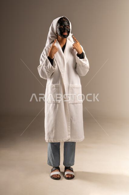 النظر الى الكاميرا بإيماءات السرور، خطوات تنظيف وتقشير البشرة، استخدام مكون ومستحضر تجميلي، بورتريه لامرأة عربية خليجية سعودية ترتدي منشفة وروب الاستحمام الابيض تضع قناع الطين تشير الى وجهها باصبعي السبابة، صورة الجسم بالطول الكامل، خلفية رمادية