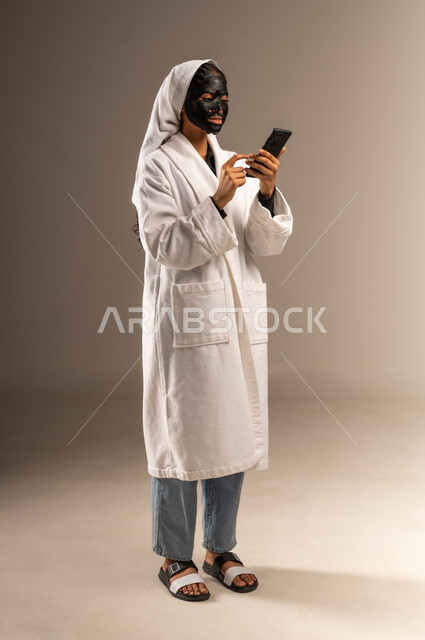 خطوات تنظيف وتقشير البشرة، استخدام مكون و مستحضر تجميلي، بورتريه لامرأة عربية خليجية سعودية ترتدي منشفة وروب الاستحمام الابيض تضع قناع الطين على وجهها تقوم باستخدام الهاتف المحمول، الدردشة عبر مواقع التواصل الاجتماعي، صورة الجسم بالكامل، خلفية رمادية