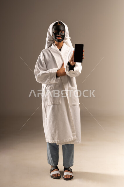 استخدام مستحضر تجميلي, عرض شاشة سوداء فارغه عبر الهاتف المحمول, خطوات تنظيف وتقشير البشرة، بورتريه لامرأة عربية خليجية سعودية ترتدي منشفة وروب الاستحمام الابيض تضع قناع الطين على وجهها تنظر الى الكاميرا و تمسك بيدها الجوال، صورة الجسم بالكامل، خلفية رمادية