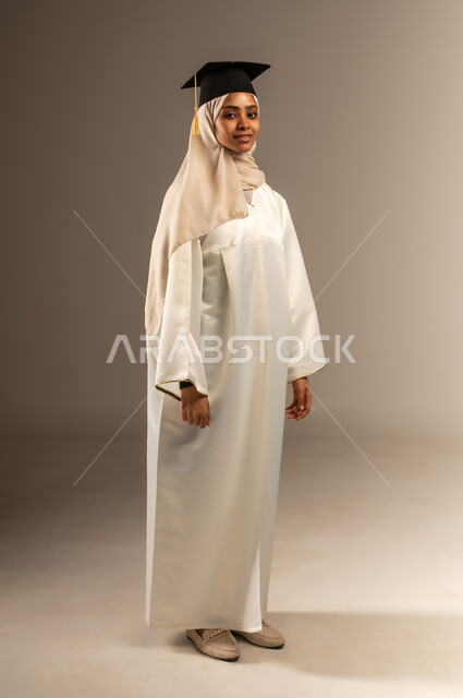 الوقوف باستقامة و النظر الى الكاميرا بإيماءات الفخر والثقة بالنفس، الأناقة والاهتمام بالمظهر الخارجي, المناسبات والاحتفالات الجامعية, بورتريه لطالبة عربية سعودية خليجية محجبة ترتدي ثوب التخرج الابيض و القبعة, صورة الجسم بالطول الكامل, خلفية رمادية