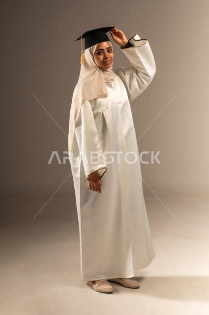 الأناقة والاهتمام بالمظهر الخارجي, المناسبات والاحتفالات الجامعية, بورتريه لطالبة عربية سعودية خليجية محجبة ترتدي ثوب التخرج الابيض تمسك بيدها القبعة تنظر الى الكاميرا بإيماءات الفخر والثقة بالنفس, صورة الجسم بالطول الكامل, خلفية رمادية