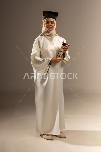 الاهتمام بالأناقة والمظهر الخارجي, المناسبات والاحتفالات الجامعية, بورتريه لطالبة عربية سعودية خليجية محجبة ترتدي ثوب التخرج الابيض والقبعة تمسك بيدها الوثيقة تنظر الى الكاميرا بإيماءات الفخر, صورة الجسم بالطول الكامل, خلفية رمادية
