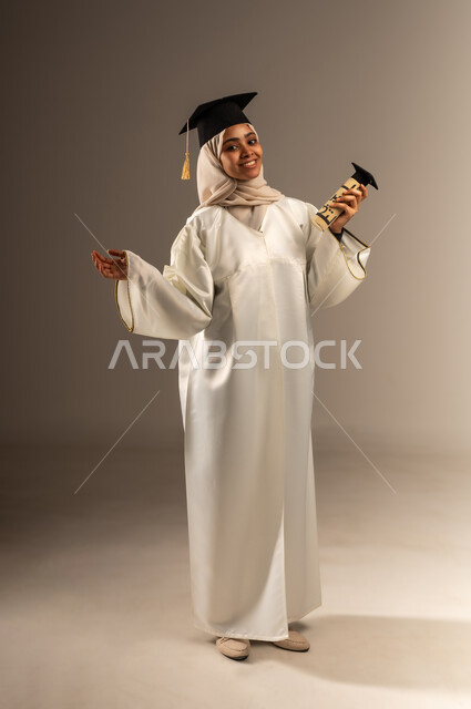 الشعور بالسرور و الفرح لتحقيق النجاح, المناسبات والاحتفالات الجامعية, بورتريه لطالبة عربية سعودية خليجية محجبة ترتدي ثوب التخرج الابيض والقبعة تنظر الى الكاميرا ترفع يديها للاعلى تمسك الوثيقة بإيماءات الفخر, صورة الجسم بالطول الكامل, خلفية رمادية