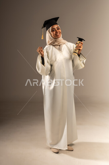 تعابير السرور و الفرح لتحقيق النجاح, المناسبات والاحتفالات الجامعية, بورتريه لطالبة عربية سعودية خليجية محجبة ترتدي ثوب التخرج الابيض والقبعة تنظر الى الكاميرا ترفع يديها للاعلى تمسك الوثيقة بإيماءات الفخر, صورة الجسم بالطول الكامل, خلفية رمادية