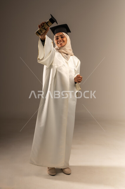Feeling happy and joyful for achieving success, university occasions and celebrations, portrait of a veiled Saudi Arabian Gulf female student wearing a white graduation gown and cap looking at the camera raising her hands up holding the document with gestures of pride, full body photo, gray background