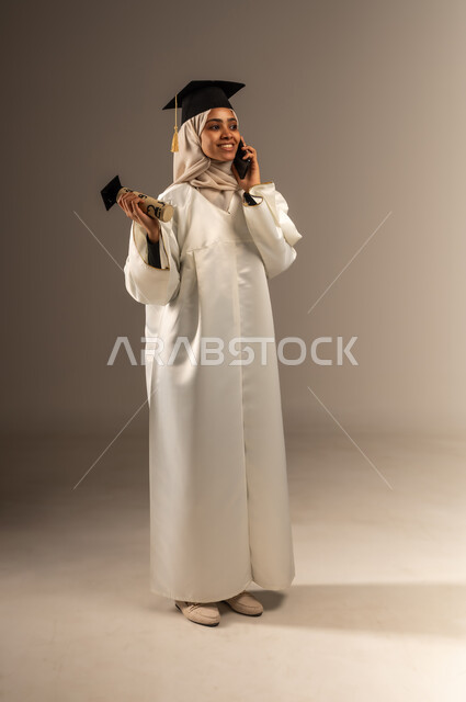 التواصل مع العائلة و الأصدقاء، استخدام جهاز تقني حديث, المناسبات والاحتفالات الجامعية, بورتريه لطالبة عربية سعودية خليجية محجبة ترتدي ثوب التخرج الابيض والقبعة تمسك بيدها الوثيقة والهاتف المحمول تقوم بإجراء مكالمة هاتفية, صورة الجسم بالكامل, خلفية رمادية
