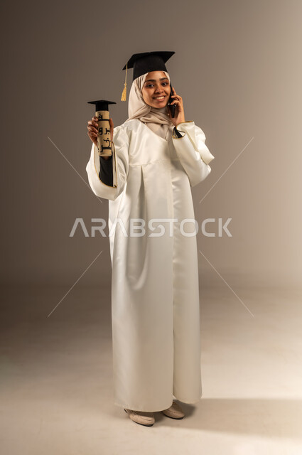 Using a modern technical device, university events and celebrations, portrait of a veiled Saudi Arabian Gulf female student wearing a white graduation gown and hat holding a document and a mobile phone in her hand, looking at the camera, making a phone call, full body image, gray background
