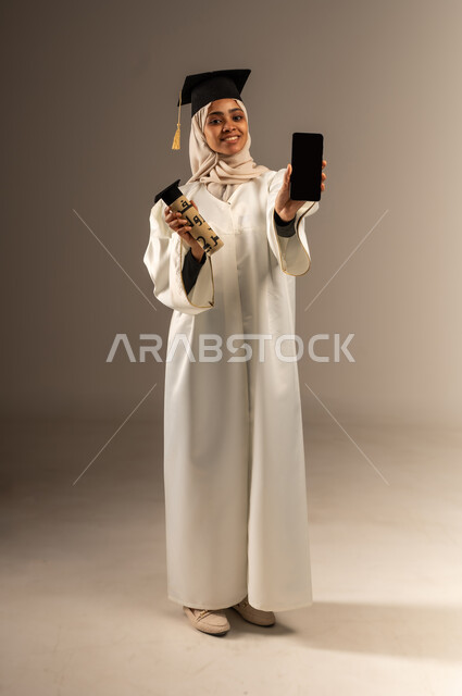 عرض شاشة سوداء فارغة عبر الهاتف المحمول, استخدام جهاز تقني حديث, أهمية الدراسة الجامعية, بورتريه لشابة عربية سعودية خليجية محجبة ترتدي القبعة وعباءة التخرج تنظر الى الكاميرا بايماءات السعادة تمسك بيدها شهادة التخرج, صورة الجسم بالطول الكامل, خلفية رمادية