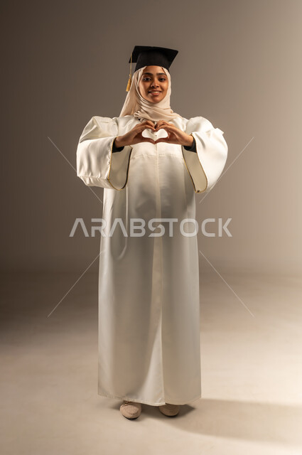 University events and celebrations, feeling happy and joyful for achieving success, portrait of a veiled Saudi Arabian Gulf student wearing a white graduation gown and hat looking at the camera raising her hands up in the shape of a heart, higher education in the Kingdom, full-length body image, gray background
