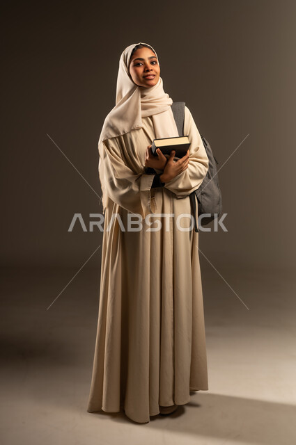 الاستعداد للعام الدراسي الجديد, التعليم العالي في الجامعات الأكاديمية في السعودية, بورتريه لطالبة جامعية عربية خليجية سعودية مبتسمة محجبة ترتدي العباءة تضع حقيبة الظهر تحمل بيدها كتاب تنظر الى الكاميرا بإيماءات السرور, صورة الجسم بالطول الكامل، خلفية رمادية