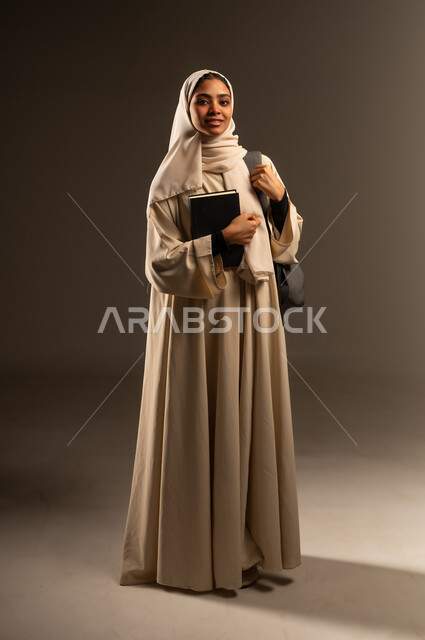 التعليم العالي في الجامعات الأكاديمية في السعودية, الاستعداد للعام الدراسي الجديد, بورتريه لطالبة جامعية عربية خليجية سعودية مبتسمة محجبة ترتدي العباءة تضع حقيبة الظهر تحمل بيدها كتاب تنظر الى الكاميرا بإيماءات السرور, صورة الجسم بالطول الكامل، خلفية رمادية