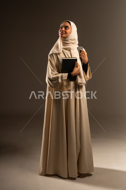 الاستعداد للعام الدراسي الجديد, تعابير النشاط والاجتهاد، التعليم العالي في الجامعات الأكاديمية في السعودية, بورتريه لطالبة جامعية عربية خليجية سعودية مبتسمة محجبة ترتدي العباءة تضع حقيبة الظهر تحمل بيدها كتاب تنظر الى شيء ما في الأعلى, صورة الجسم بالكامل، خلفية رمادية