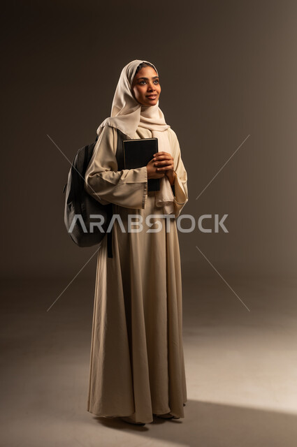 Expressions of activity and diligence, higher education in academic universities in Saudi Arabia, portrait of a smiling Saudi Arabian Gulf university student wearing a hijab, wearing an abaya and putting a backpack holding a book in her hand, preparing for the new school year, full-length body image, gray background