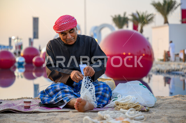 أدوات ومعدات صيد السمك, فعالية ونشاط ثقافي تراثي, صياد عربي خليجي قطري يرتدي الثوب التقليدي والشماغ يقوم بتحضير شبكة الصيد, الاحتفال بمهرجان كتارا للمحامل التقليدية على شاطئ مدينة الدوحة, مهن وأعمال شعبية