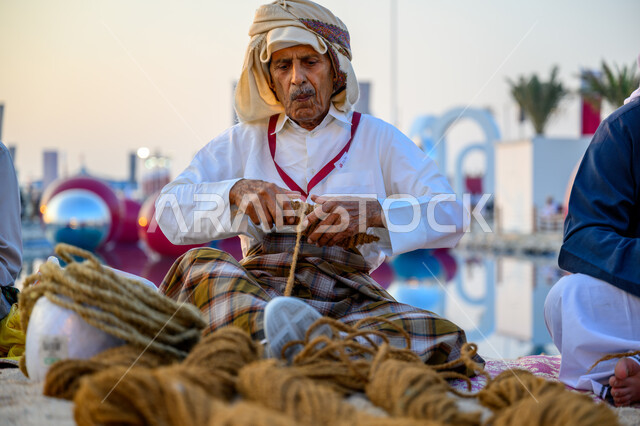 Cultural heritage event and activity, fishing tools and equipment, a Qatari Gulf Arab fisherman wearing traditional dress preparing a fishing net, celebrating the Katara Traditional Dhow Festival on the beach of Doha, popular professions and businesses