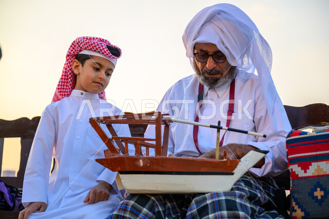 تراث الأجداد القطري, مهرجان كتارا للقوارب الشراعية التقليدية, الاحتفال بالأعياد والمناسبات الوطنية, ذكرى باليوم الوطني لدولة قطر 18 ديسمبر, فعالية ونشاط ترفيهي احتفالي, صياد عربي خليجي قطري يرتدي الثوب التقليدي يجلس مع ابنه ويمسك بيده مجسم قارب صيد خشبي صغير