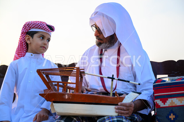 مهرجان كتارا للقوارب الشراعية التقليدية, تراث الأجداد القطري, الاحتفال بالأعياد والمناسبات الوطنية, ذكرى باليوم الوطني لدولة قطر 18 ديسمبر, فعالية ونشاط ترفيهي احتفالي, صياد عربي خليجي قطري يرتدي الثوب التقليدي يجلس مع ابنه ويمسك بيده مجسم قارب صيد خشبي صغير