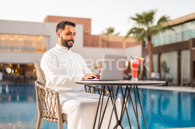 تناول عصير البطيخ المنعش في الهواء الطلق، استخدام التقنيات الحديثة والتكنولوجيا بالكافيهات والمقاهي، إنجاز مهام العمل عن بعد عبر الحاسوب المحمول، رجل عربي خليجي سعودي يرتدي الثوب التقليدي ويجلس على طاولة حول المسبح في أحد المطاعم في المملكة العربية السعودية
