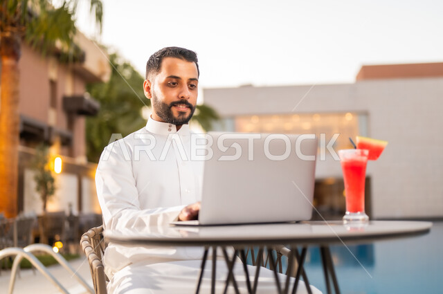 استخدام التقنيات الحديثة والتكنولوجيا بالكافيهات والمقاهي، إنجاز مهام العمل عن بعد عبر الحاسوب المحمول، رجل عربي خليجي سعودي يرتدي الثوب التقليدي ويجلس على طاولة حول المسبح في أحد المطاعم في المملكة العربية السعودية، تناول عصير البطيخ المنعش في الهواء الطلق