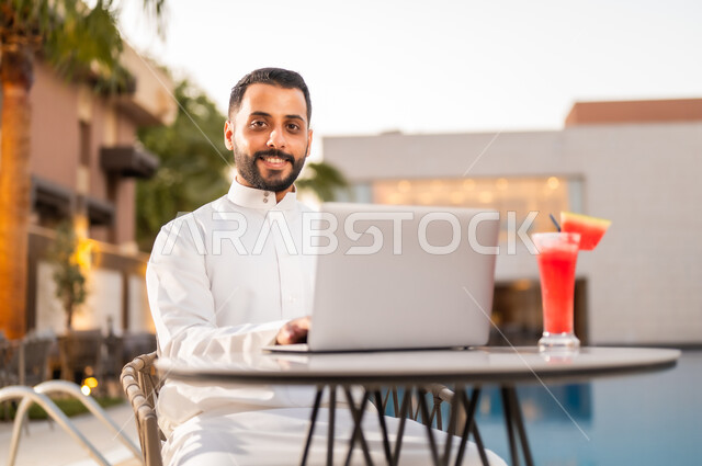 إنجاز مهام العمل عن بعد عبر الحاسوب المحمول، النظر إلى الكاميرا بإيماءات الرضا والسعادة، رجل عربي خليجي سعودي يرتدي الثوب التقليدي ويجلس على طاولة حول المسبح في أحد المطاعم في المملكة العربية السعودية، استخدام التقنيات الحديثة والتكنولوجيا بالكافيهات والمقاهي