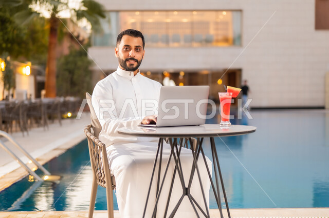 النظر إلى الكاميرا بإيماءات الرضا والسعادة، رجل عربي خليجي سعودي يرتدي الثوب التقليدي ويجلس على طاولة حول المسبح في أحد المطاعم في المملكة العربية السعودية، إنجاز مهام العمل عن بعد عبر الحاسوب المحمول، استخدام التقنيات الحديثة والتكنولوجيا بالكافيهات والمقاهي