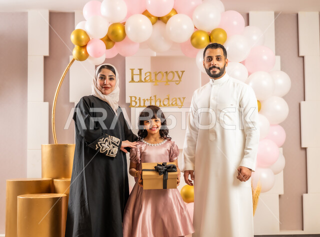 النظر إلى الكاميرا بإيماءات السعادة والسرور بجو عائلي دافئ، التزيين وتعليق البلالين والاحتفال في أحد المطاعم الفاخرة في المملكة، أجواء سعيدة لأسرة سعودية، زوجان عربيان خليجيان سعوديان يرتديان الملابس التقليدية يقومان بالاحتفال بعيد ميلاد ابنتهما الصغيرة