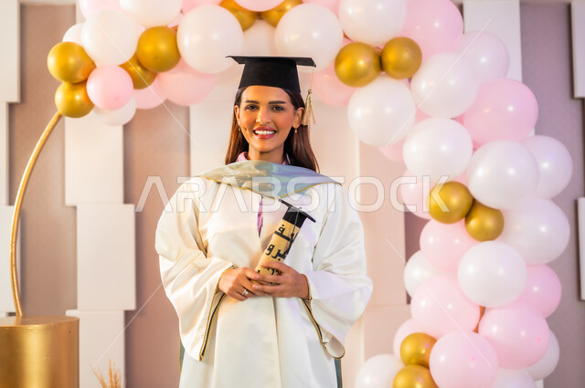 النظر إلى الكاميرا بإيماءات السعادة والسرور بتحقيق الأحلام المنتظرة، الفخر بالنجاح بعد مواجهة التحديات والتعب, طالبة جامعية عربية خليجية سعودية ترتدي قبعة وثوب التخرج الأبيض وتمسك الوثيقة بمشاعر الفرح والامتنان, تعليق البالونات والزينة في المناسبات السعيدة