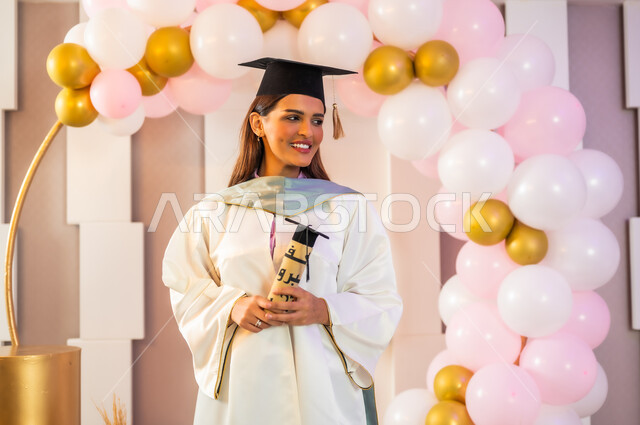 الاحتفال بالوصول إلى الأحلام وتحقيقها، إيماءات الفخر والسرور بالنجاح بعد مواجهة التحديات والتعب, طالبة جامعية عربية خليجية سعودية ترتدي قبعة وثوب التخرج الأبيض وتمسك الوثيقة بمشاعر الفرح والامتنان, التزيين وتعليق البالونات في الاحتفالات والمناسبات السعيدة