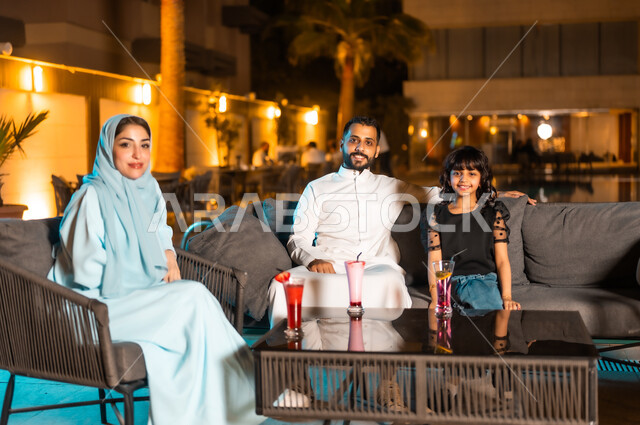 الخروج ضمن أجواء عائلية لطيفة، القيام بنشاطات ممتعة بعطلة نهاية الأسبوع، الاستمتاع بالمشروبات الباردة في المطاعم والمقاهي بالمملكة العربية السعودية، رجل عربي خليجي سعودي يرتدي الثوب التقليدي برفقة طفلته وزوجته ينظرون إلى الكاميرا بتعابير السعادة والسرور