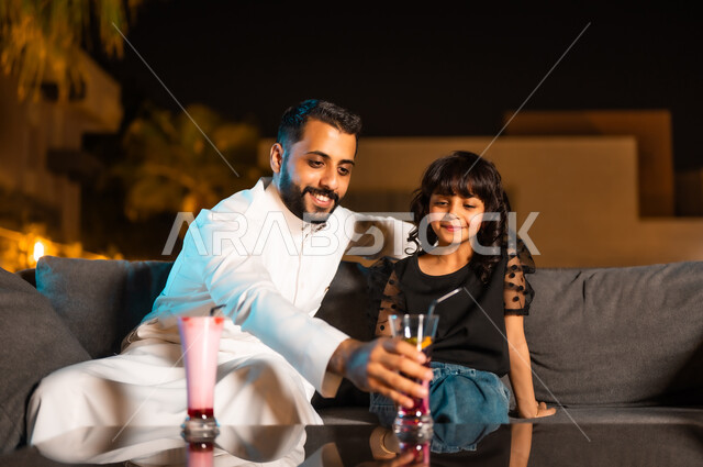 الاستمتاع بعطلة نهاية الأسبوع والقيام بنشاطات برفقة العائلة، تقوية روابط المودة والحب بين الأب وابنته، أجواء صيفية لطيفة في المطاعم والمقاهي بالمملكة العربية السعودية، رجل عربي خليجي سعودي يرتدي الثوب التقليدي برفقة طفلته يتناولان العصائر الباردة المنعشة