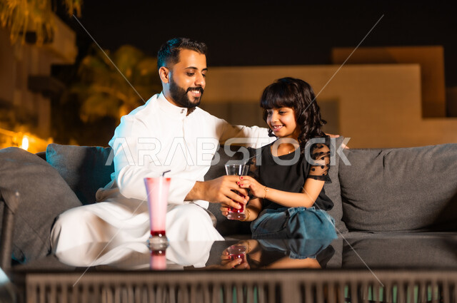 القيام بنشاطات ممتعة برفقة العائلة في عطلة نهاية الأسبوع، تقوية روابط المودة والحب بين الأب وابنته، أجواء صيفية لطيفة في المطاعم والمقاهي بالمملكة العربية السعودية، رجل عربي خليجي سعودي يرتدي الثوب التقليدي برفقة طفلته يتناولان العصائر الباردة المنعشة